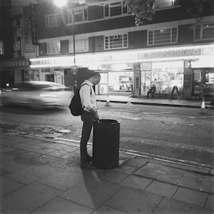 May include: Black and white street scene at night. A person stands next to a dark trash can, appearing to urinate. The background includes a storefront with illuminated signs and a blurred car.