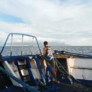 Puede incluir: Una persona está de pie en la cubierta de un barco, mirando al mar. El barco está pintado de azul y blanco, con cuerdas y barandillas azules. La persona lleva una camiseta sin mangas con estampado en blanco y negro y pantalones cortos color caqui. El cielo está nublado.