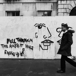 May include: Black and white photo of a person walking past a white wall with black graffiti. The graffiti includes a face and the words "Fell through the rabbit hole ended up here".