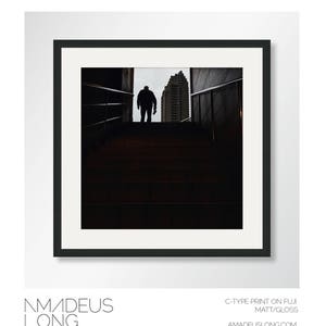 Puede incluir: Fotograf&iacute;a en blanco y negro enmarcada que presenta una figura silueteada en la parte superior de una escalera, con un edificio al fondo. La imagen es una impresi&oacute;n C-type sobre papel Fuji Matt/Gloss. El nombre del artista, Amadeus Long, est&aacute; en la parte inferior.