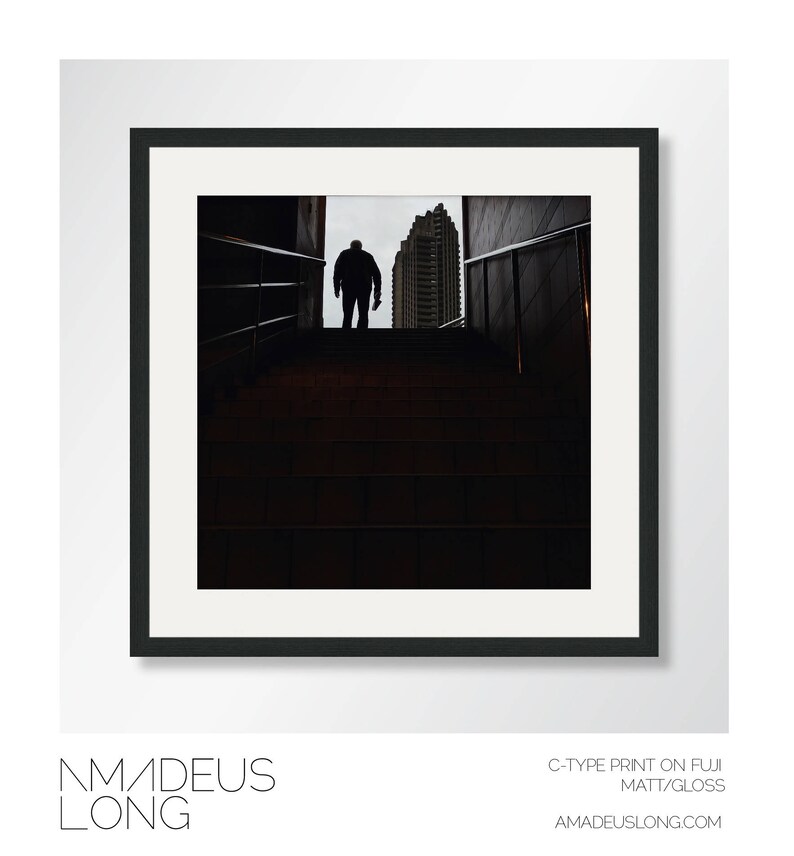 Puede incluir: Fotograf&iacute;a en blanco y negro enmarcada que presenta una figura silueteada en la parte superior de una escalera, con un edificio al fondo. La imagen es una impresi&oacute;n C-type sobre papel Fuji Matt/Gloss. El nombre del artista, Amadeus Long, est&aacute; en la parte inferior.