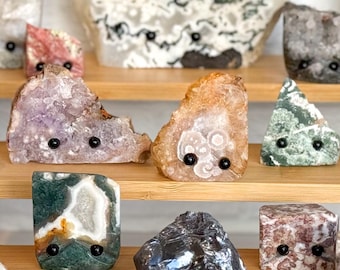 Puede incluir: Una colección de varios especímenes minerales en bruto dispuestos en un expositor de madera de varios niveles. Cada piedra presenta dos pequeños ojos negros redondos fijados en la parte frontal, dándoles una apariencia de personaje con texturas variadas.