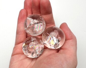 May include: Three clear quartz crystal spheres with iridescent rainbow colours inside. The spheres are in a person's hand.