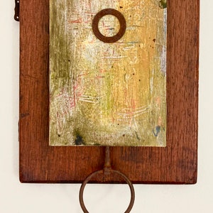 May include: A rustic wooden wall hanging with a painted panel featuring a circular cutout and a rusty metal hook with two rings hanging below.