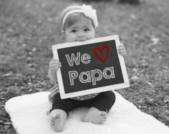 May include: A baby holding a black chalkboard with the words "We ♥ Papa" written in white chalk. The heart is drawn in red.