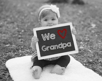 Könnte beinhalten: Ein kleines Kind hält eine schwarze Tafel mit den Worten "We ♥ Grandpa" in weißer Kreide geschrieben. Das Kind trägt ein weißes Stirnband mit einer Blume und ein weiß-schwarz gemustertes Kleid.