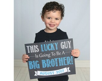 May include: A young boy holds a black chalkboard sign that says "This Lucky Guy Is Going To Be A Big Brother! August 2017". The sign has white and blue lettering.