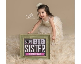 May include: A young girl in a cream-colored dress stands near a framed sign. The sign reads "FUTURE BIG SISTER AUGUST 2018" in white and pink lettering against a black background, framed in green. The girl is smiling and looking at the sign.