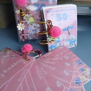 May include: Pink floral mini ring binder notebooks with gold rings. One notebook has a pink pom-pom charm and bee charms. A fan of pink, labelled cards with white text is also visible.