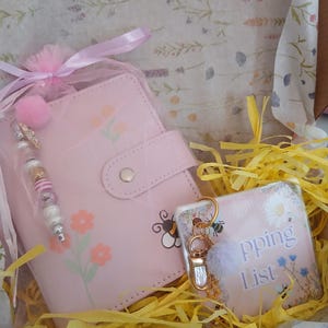 May include: A light pink notebook with a snap closure, decorated with orange flowers and a bee illustration, accompanied by a beaded charm. Next to it is a square shopping list pad featuring a bee design and a gold-toned keychain with a pom-pom.