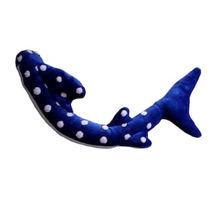 May include: A plush whale shark toy featuring a deep blue body covered in white polka dots. The soft stuffed animal has a curved, elongated shape with a prominent dorsal fin, smaller secondary fins, and a wide, forked tail fin, with a white underside.