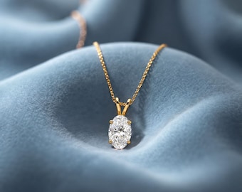 May include: A gold necklace featuring a single oval-cut clear gemstone pendant held by a four-prong setting. The delicate gold chain connects to the pendant, which rests centered on the fabric. The design is simple, focusing on the stone.