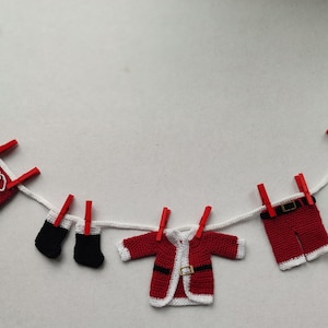 May include: A string of miniature crocheted Santa Claus outfits hanging on a clothesline with red clothes pegs. The outfits include a hat, jacket, trousers, and boots.