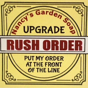 May include: A yellow and black sign with the text "Nancy's Garden Soap" in a circle at the top. The text "UPGRADE" is below the circle. A white banner with the text "RUSH ORDER" in red letters is below the text "UPGRADE". The text "PUT MY ORDER AT THE FRONT OF THE LINE" is below the banner.