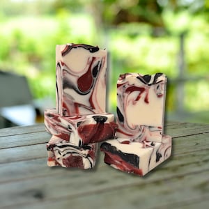 May include: A stack of handmade soap bars with a marbled design. The soap features swirls of white, black, and burgundy colors. The bars are rectangular and have a rustic, natural appearance, suggesting a focus on artisanal craftsmanship.