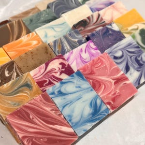 May include: A collection of rectangular soap bars arranged in a grid, each featuring unique marbled patterns in various colors including pink, blue, purple, orange, brown, and white. The soaps have smooth surfaces with swirling designs.