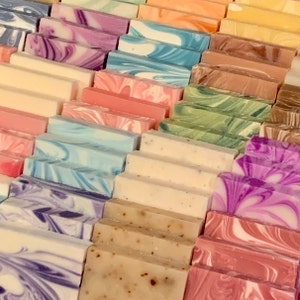 May include: A collection of rectangular soap bars arranged in rows. The bars feature various designs, including solid colors, speckled textures, and swirling patterns in shades of blue, purple, pink, green, brown, orange, and cream.