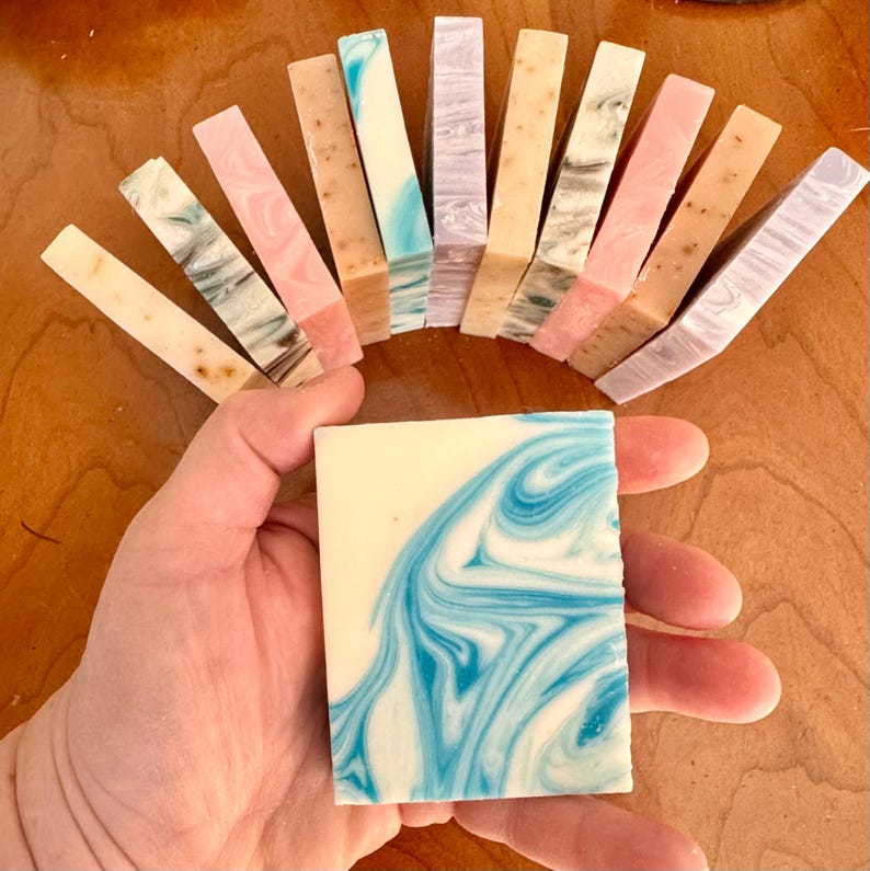 May include: A collection of handmade soap bars in various colors and patterns, including blue, pink, and brown. The bars are arranged in a semi-circle, with a single bar in the center of the image.