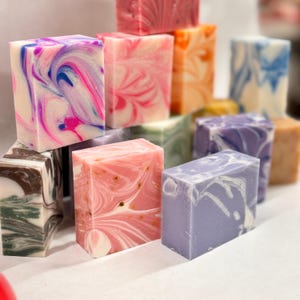 May include: A collection of handmade soap bars in various colors and patterns, including pink, purple, blue, and brown. The soap bars are arranged in a pyramid shape.