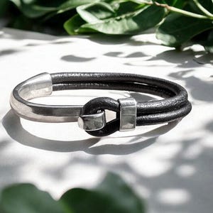 May include: A bracelet featuring two strands of black leather cord secured by a polished silver-toned metal hook clasp. The design incorporates a smooth, curved silver-toned metal bar that connects to the leather cords, creating a continuous loop.