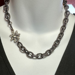 Puede incluir: Un collar de cadena negra con un colgante de flor de pedrería plateada.