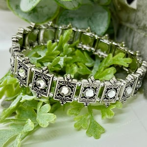 Op de afbeelding: Een zilveren stretch armband met heldere strass accenten in een vierkante vorm.