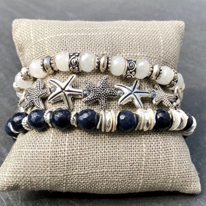 May include: A set of four bracelets featuring white and blue beads, silver starfish charms, and silver spacer beads. The bracelets are displayed on a gray fabric surface.