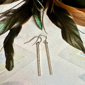 May include: A pair of gold dangle earrings with a long, thin bar design. The bars are encrusted with small, clear rhinestones.