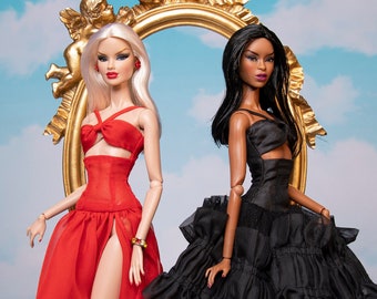 May include: Two fashion dolls stand before a gold-colored ornate mirror against a sky-blue backdrop. One doll wears a red gown with a high slit, the other a black dress with tiered ruffles. Both dolls have high heels.