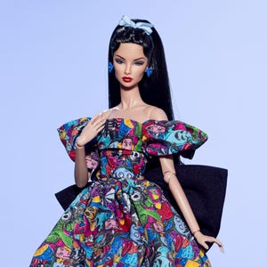May include: A fashion doll wearing a colorful off-the-shoulder gown with a black bow. The gown features a vibrant pattern of cartoon faces in various colors. The doll has long black hair, blue earrings, and a matching bow.