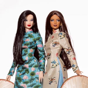 May include: Two dolls wearing traditional Vietnamese áo dài dresses and conical hats. One doll's dress is light blue with a lotus flower pattern, while the other is beige with colorful embroidered designs. Both dolls have long, dark hair.