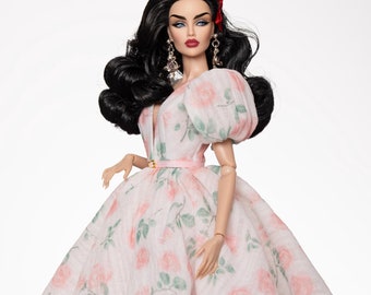 May include: A doll wearing a white dress with a pink floral pattern and a matching belt. The doll has long black hair and is wearing red earrings.