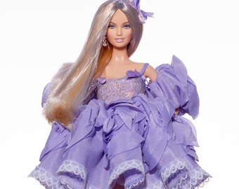May include: A fashion doll with long blonde hair and a lavender dress. The dress features ruffled layers, lace trim, and a sparkly bodice. The doll wears gold-colored heels and a matching hair accessory. The doll is standing against a white background.