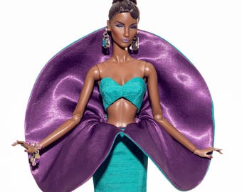 May include: A fashion doll in a teal and purple gown. The doll has dark skin, brown hair, and is wearing large earrings and a bracelet. The gown features a teal bodice and a flared skirt, with a large purple accent around the shoulders.