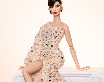 May include: A fashion doll wearing a cream-colored, one-shoulder gown adorned with gold sequins. The doll is seated on a white, decorative column, wearing red heels and gold earrings. The doll has dark hair and red lipstick.