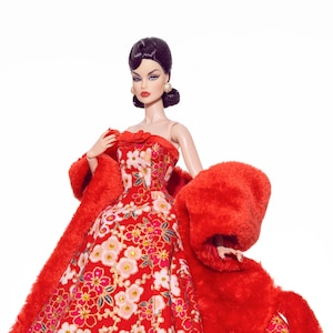 Könnte beinhalten: Modepuppe in einem roten Kleid mit Blumenmuster und passendem roten Kunstfell-Wrap. Das trägerlose Kleid hat einen weiten Rock und ist mit weißen und rosa Blumen verziert. Die Puppe hat dunkles Haar und trägt goldene Ohrringe.