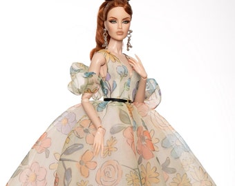 Puede incluir: Una muñeca con un vestido blanco con estampado floral y un cinturón negro. La muñeca tiene el pelo largo y rojo y lleva zapatos marrones.