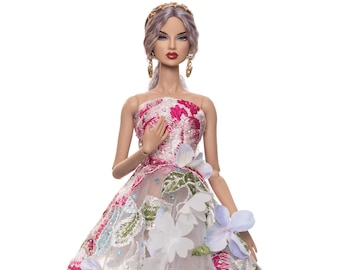 Puede incluir: Una muñeca con un vestido blanco con bordado floral y zapatos de color morado claro. El vestido tiene flores rosas, azules y verdes.