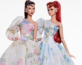 May include: Two fashion dolls in floral, one-shoulder dresses with full skirts and waist belts. One doll has dark hair, the other red. Both wear high heels. The dresses are in a pastel floral print.