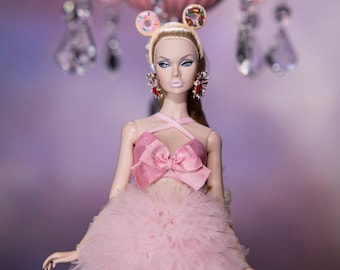 Puede incluir: Una muñeca rosa y blanca con una falda de tul rosa y un top de lazo rosa. La muñeca lleva una diadema de donut y pendientes. La muñeca está de pie frente a una lámpara de araña rosa.