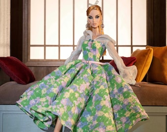 May include: A doll wearing a green and white floral dress with a white sheer overlay and a pink ribbon at the waist. The doll has blonde hair and is wearing earrings. The doll is sitting on a sofa with cushions.