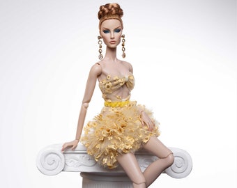 May include: A fashion doll with long, auburn hair styled in a bun, wearing a yellow and gold tulle dress with floral embellishments. The doll is sitting on a white pedestal with her legs crossed and wearing clear high heels.