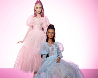 Puede incluir: Dos muñecas de moda con vestidos de tul elaborados. La muñeca de la izquierda lleva un vestido rosa con mangas abullonadas y la muñeca de la derecha lleva un vestido azul claro con mangas abullonadas. Ambas muñecas llevan pendientes.