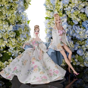 May include: Two dolls wearing floral print dresses and sitting in a floral setting. One doll is wearing a long white dress with a floral pattern and the other doll is wearing a white dress with a floral pattern and a white jacket.
