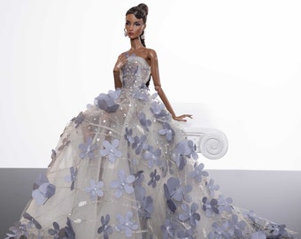 May include: A fashion doll wearing a strapless gown with a tulle skirt and blue flower embellishments. The doll has dark brown hair and brown eyes.