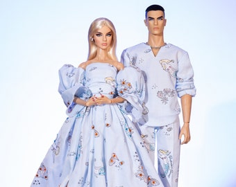 Puede incluir: Un par de muñecas que llevan conjuntos a juego de color azul claro con un estampado de sirena y vida marina. La muñeca de la izquierda lleva un vestido largo y fluido con un escote de volantes. La muñeca de la derecha lleva una camisa con cuello y pantalones. Ambas muñecas tienen el pelo rubio y están de pie sobre un fondo blanco.