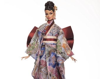 Puede incluir: Muñeca de moda con un kimono colorido con diseños de abanicos. El kimono presenta tonos de azul, rosa y verde, con un gran lazo burdeos en la espalda. La muñeca tiene el pelo oscuro recogido en un moño y lleva pendientes dorados.