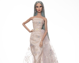 May include: A fashion doll wearing a white strapless gown with a sheer overlay and intricate embroidery. The doll has long, flowing blue hair and is wearing delicate earrings.