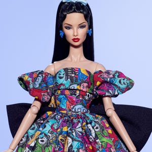 May include: A fashion doll with long black hair, red lipstick, and blue earrings and bow. The doll wears a colorful strapless dress with a black bow in the back. The dress features a print with various cartoon characters in multiple colors.
