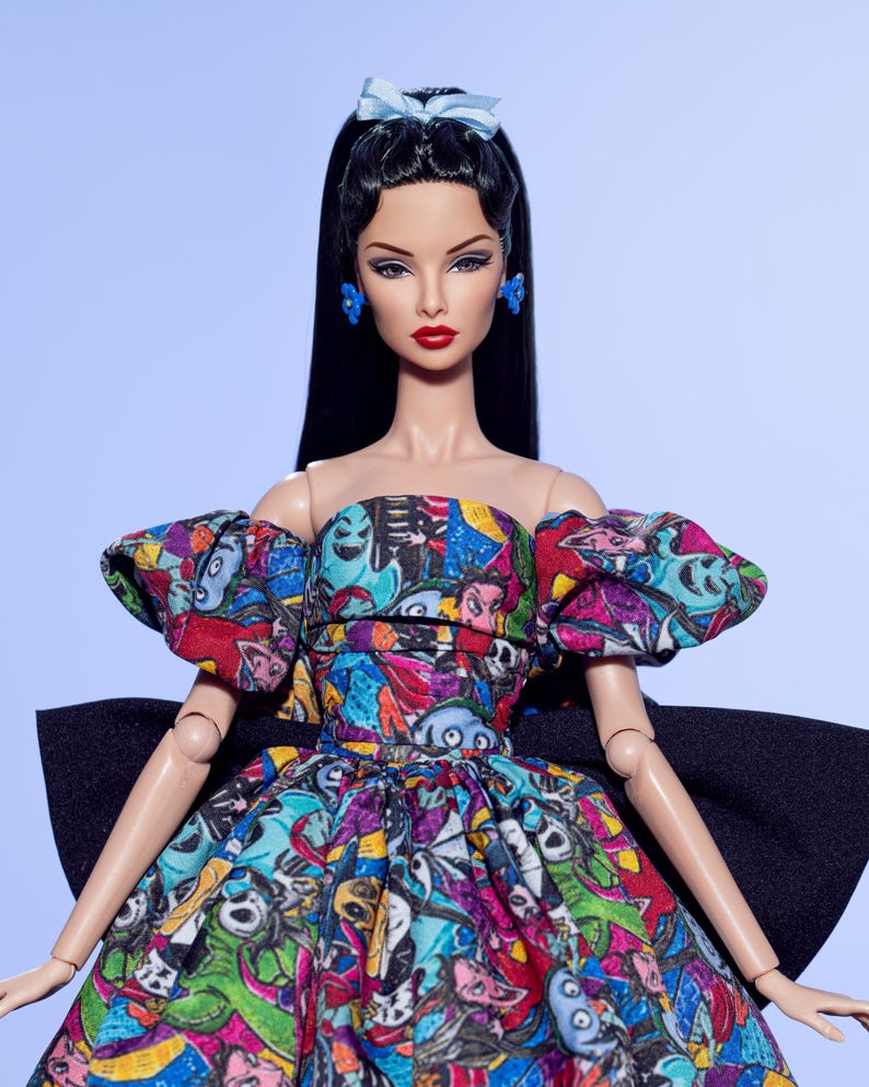 May include: A fashion doll with long black hair, red lipstick, and blue earrings and bow. The doll wears a colorful strapless dress with a black bow in the back. The dress features a print with various cartoon characters in multiple colors.